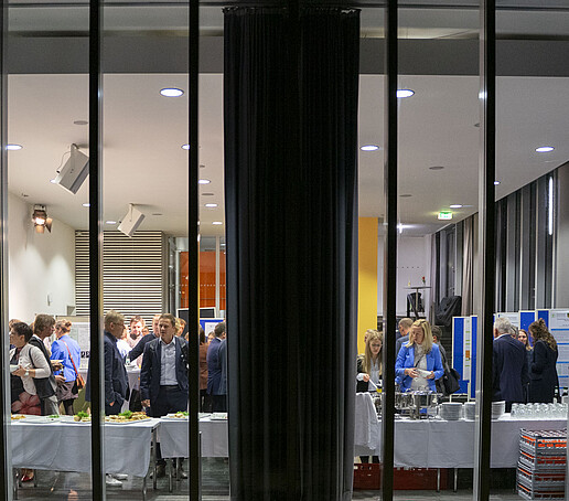 Durch die Glasscheibe von außen, sieht man die Gäste in der Aula, wie sie sich am Buffet bedienen.