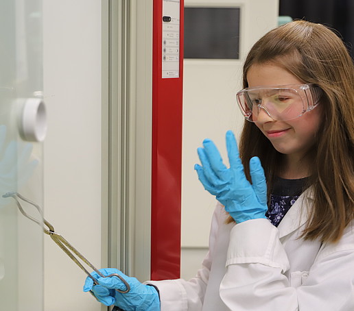 Ein Mädchen im Labor schaut fasziniert durch ihre Schutzbrille auf ihren Finger. In der anderen Hand hält sie eine Zange.