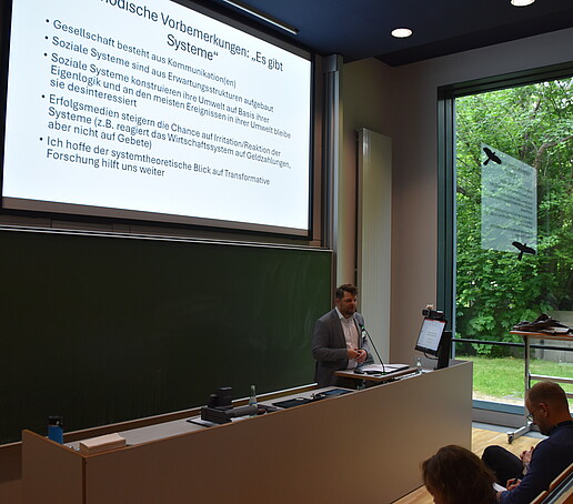 [Translate to Englisch:] 1 Redner steht am Pult im Hörsaal vor einer Präsentation