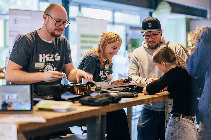 [Translate to Englisch:] 2 Mitarbeiter am Messestand erklären 2 Besuchern ein Gerät
