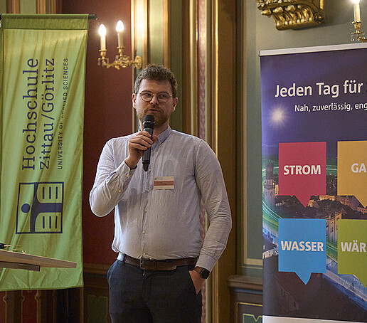 Ein Teilnehmer hält einen Vortrag. Er steht zwischen einer HSZG-Fahne und einem bunten Roll-up.