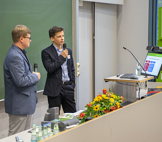 Zwei junge Wissenschaftler erläutern ihr Projekt an einer grünen Tafel.