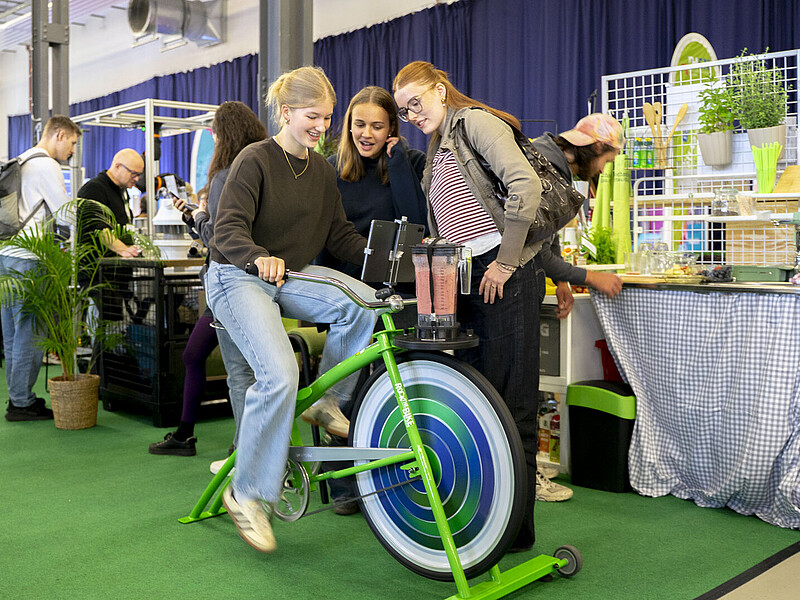 Eine Studieninteressierte radelt auf dem Smoothie-Bike, zwei Begleiterinnen stehen drumherum.
