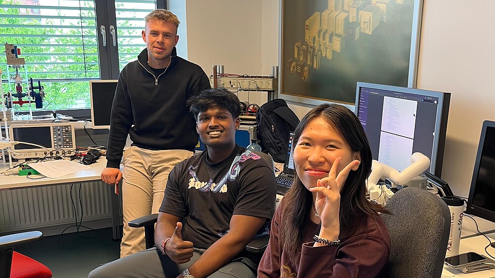Foto: HSZG Drei ausländische Studierende sitzen in einem Büro und lächeln in die Kamera.