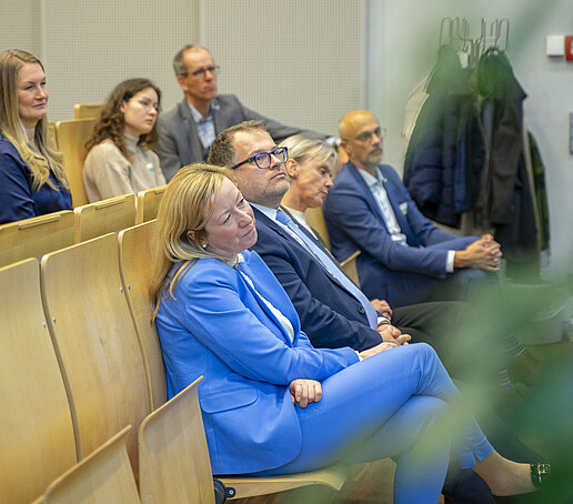 Die Sitzreihen sind mit Publikum besetzt, dass den Blick nach vorn richtet.