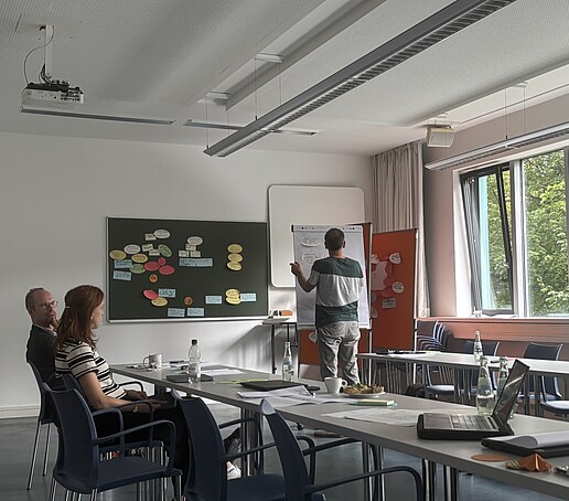 [Translate to Englisch:] 2 Teilnhemer schauen zu einem Referenten am Flipchart