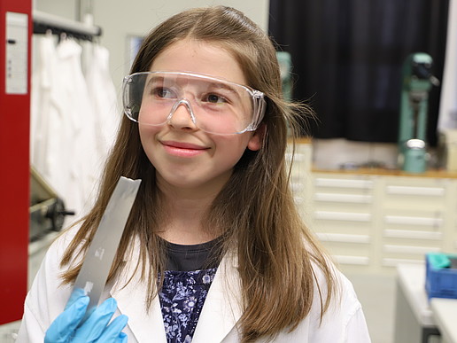 Eine Fünftklässlerin trägt einen Laborkittel und eine Schutzbrille und hält lächelnd ein Metallwerkstück in der Hand.