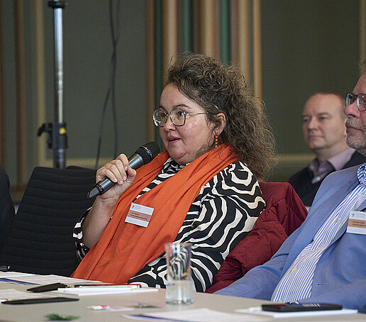 Eine Teilnehmerin mit orangenem Schal sitzt am Platz und spricht in ein Mikrofon.